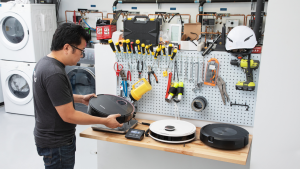 Person standing at work bench with assorted tools hanging along pegboard wall while inspecting the weight and dimensions of several small robot vacuums inside of testing lab with stacked washer and dryer combo.