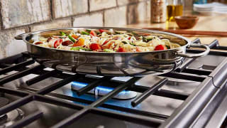 Close-up of a flavorful pasta dish in a silver pain. It sits on a gas stovetop with a blue flame glowing beneath it.