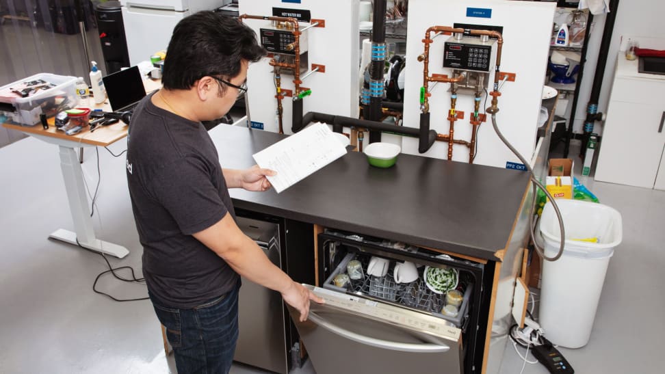 A lab tester holding a clipboard opens up a dishwasher to check on its contents