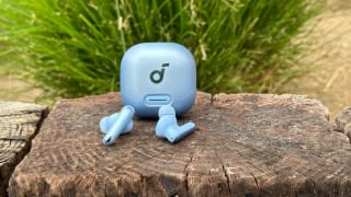 The Anker Soundcore Liberty 4 NC earbuds and case in light blue sitting on a wooden wall with a plant in the background.