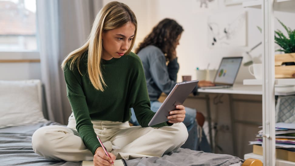 A female student sitting on a bed does homework in a dorm room while holding a tablet while another student sits at a desk behind her using a laptop.