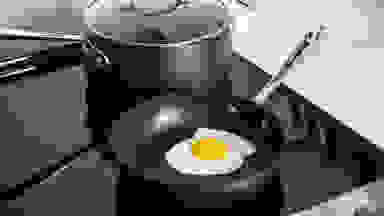 A perfect fried egg inside a nonstick pan, with a nonstick pot behind it, both sitting on a stovetop.