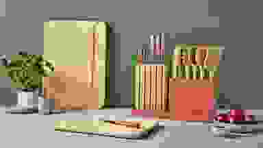 Two wooden cutting boards next to wooden cooking utensils, a knife block, a potted plant, a bowl of onions and a glass vase.