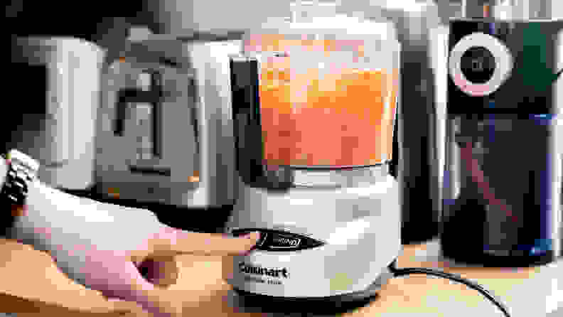 A person is pressing on the Start button of a Cuisinart mini food processor to chop carrots.