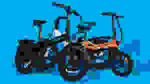 Original photograph of two folding e-bikes, one black-and-blue and one orange, against a bright blue backdrop. They have black rims and rugged black tires.