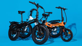 Original photograph of two folding e-bikes, one black-and-blue and one orange, against a bright blue backdrop. They have black rims and rugged black tires.