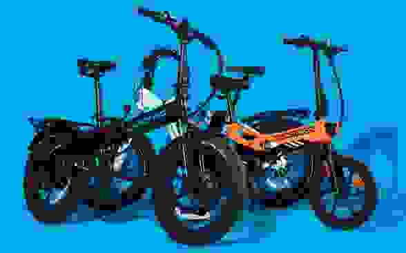 Original photograph of two folding e-bikes, one black-and-blue and one orange, against a bright blue backdrop. They have black rims and rugged black tires.
