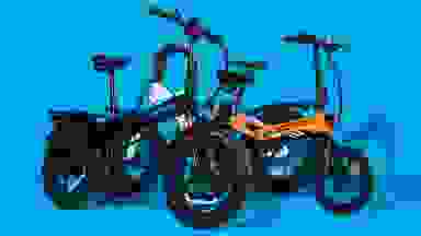 Original photograph of two folding e-bikes, one black-and-blue and one orange, against a bright blue backdrop. They have black rims and rugged black tires.