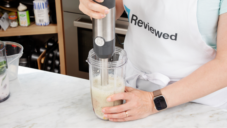 Person with apron on stands at kitchen countertop while using GE Immersion Blender to blend smoothie ingredients inside of plastic cup.