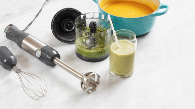 GE G8H1AASSPSS Immersion Blender next laying flat on top of granite surface next to blended green juice inside of glass, wisk attachment, soup inside of pot, and bowl.