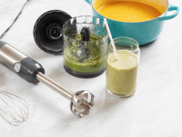 GE G8H1AASSPSS Immersion Blender next laying flat on top of granite surface next to blended green juice inside of glass, wisk attachment, soup inside of pot, and bowl.