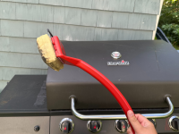 A grill brush with a foam head being held up in front of an outdoor grill.