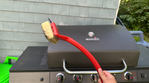 A grill brush with a foam head being held up in front of an outdoor grill.