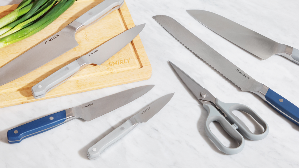 A variety of kitchen knives and a pair of sheers are spread on a marble counter and wood cutting board with scallions in the background.