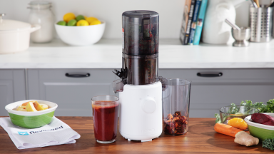 The Hurom H310A Slow Juicer sits on a wood counter in a kitchen with a full cup of juice by it, and bowls of produce on the counter.