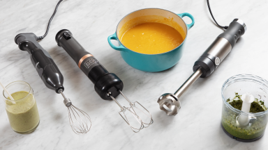 Three immersion blenders on a marble surface alongside blended soup, pesto, and a smoothie