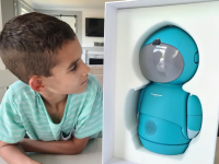 Small child smiling next to the Embodied Moxie robot in kitchen setting at table.