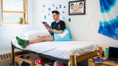 A man sits on a twin bed surrounded by shoes, school supplies, clothes, and decorations.