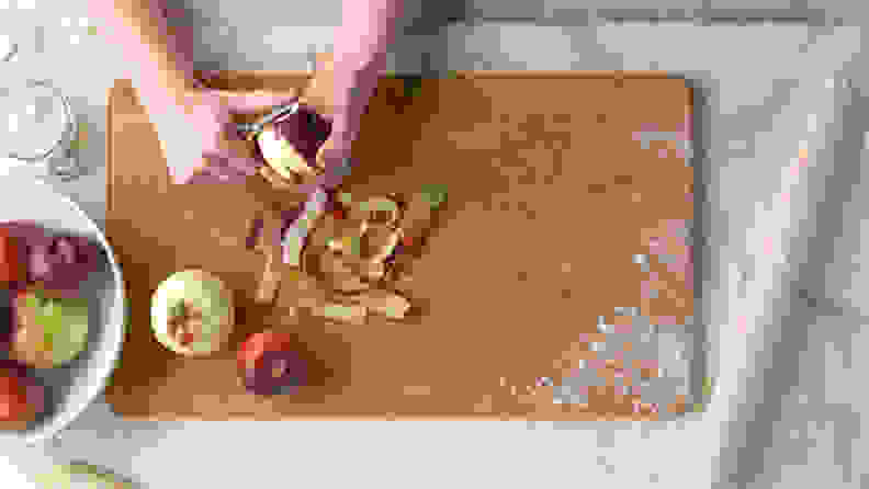 Two hand peeling an apple on a cutting board, with a rolling pin closeby