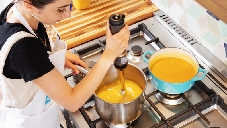 A person using the Black & Decker Kitchen Wand to blend orange sauce in a pot on a stove.