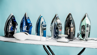 A line of steam irons sitting on top of an ironing board in front of a blue background.