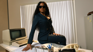 Actress Keke Palmer sits on a desk. She is wearing a black suit with dark sunglasses.