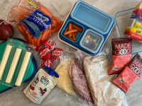 Bento boxes, cheese sticks, carrots, an apple, an assortment of deli meats and packaged snacks laid out on a marble counter.