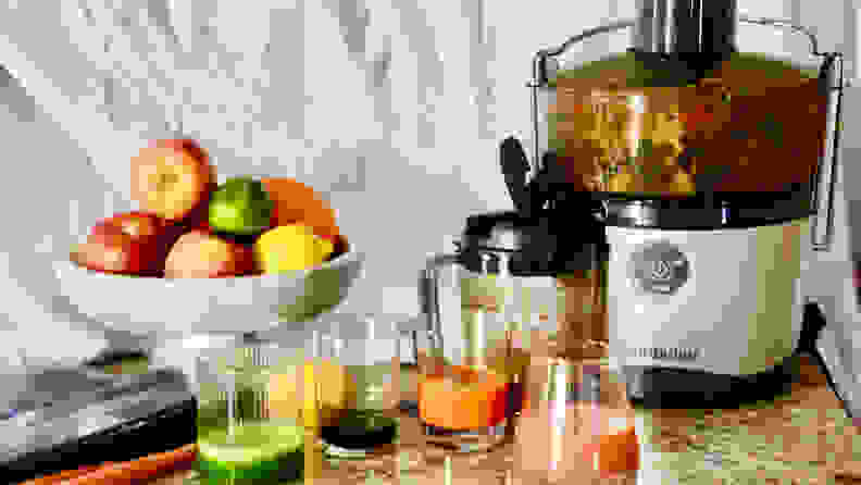 The NutriBullet Juicer Pro sits on a counter in a kitchen and is full of produce being juiced. Four glasses of different colored juices and a bowl of produce sit in the foreground.