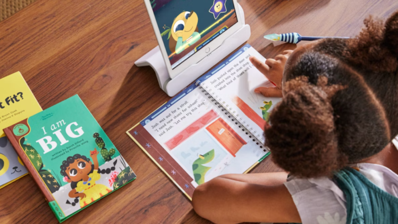 Small child sitting at table in front of tablet while reading book.