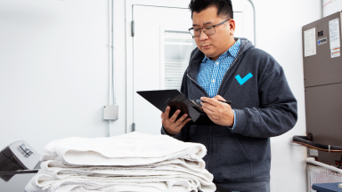 Person looks down at clipboard with pen in hand while in front of stack of white folded towels that sit on surface of black washing machine.