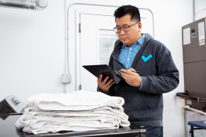 Person looks down at clipboard with pen in hand while in front of stack of white folded towels that sit on surface of black washing machine.