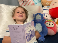 Small child smiling while laying down in bed against pillow next to Slumberkins Dragon Kin and other stuffed animals while holding book.