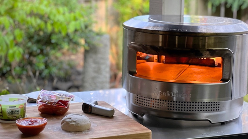 Pizza dough, marinara and pesto sauce and mozzarella cheese on wooden cutting board next to Solo Stove Pizza Oven.