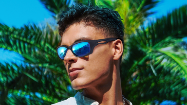 A model sports a pair of black Duduma sports sunglasses with blue mirrored lenses. A palm tree looms large in the background.