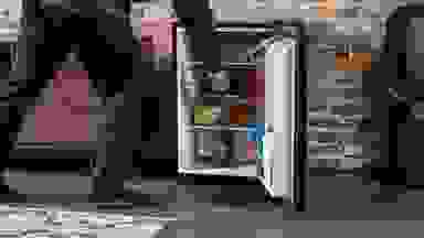 A man steps into frame to access the interior of an open Insignia NS-CF26BK9 mini fridge. It's stocked full of food and drinks.